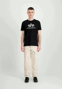 Noor mees seisab ettepoole, seljas must Alpha Industries t-särk, beežid cargo-püksid ja mustad kingad, taustal üksluiselt hele taust.