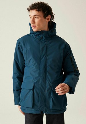 Mężczyzna w niebieskiej zimowej kurtce z kapturem i dużymi przednimi kieszeniami, patrzący w lewo na jednolitym tle.