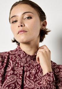 Blusa bordeaux scuro con un collo alto arricciato e maniche lunghe, caratterizzata da intricati motivi floreali rosa chiaro e polsini elasticizzati.