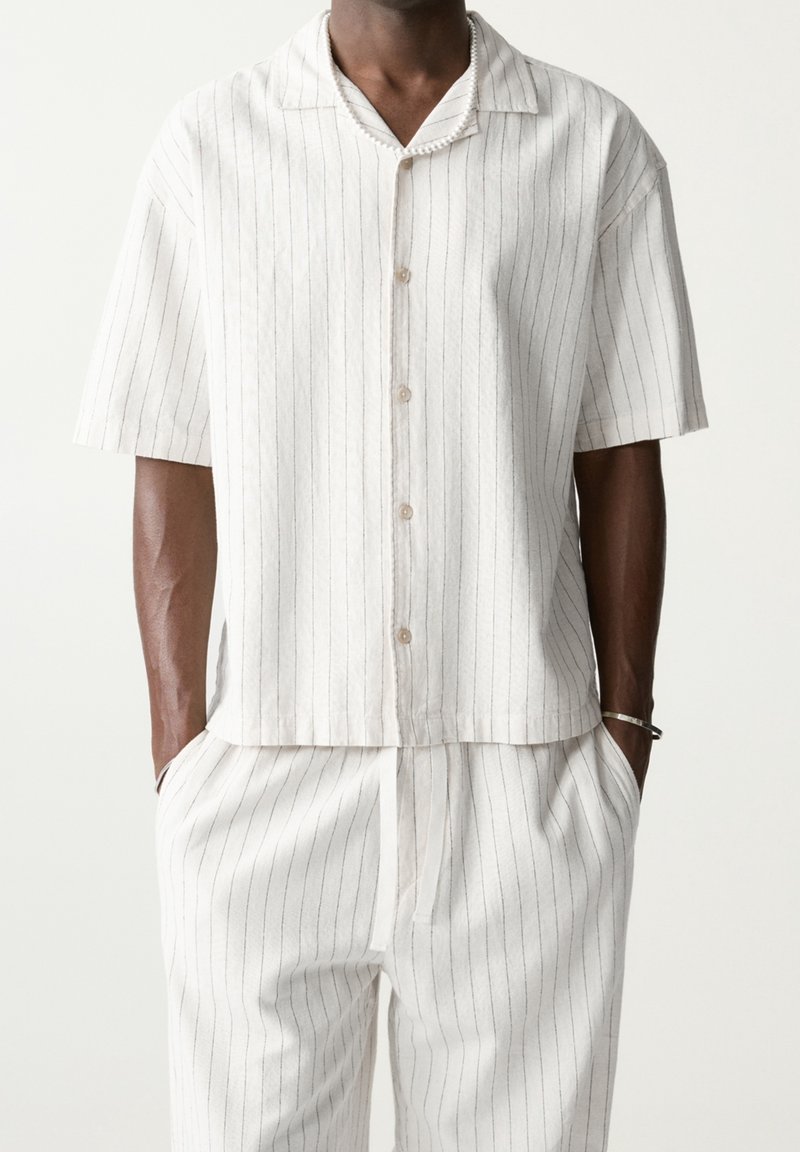 Homme portant une chemise blanche à manches courtes boutonnée et un pantalon assorti avec de fines rayures noires verticales, mains dans les poches.
