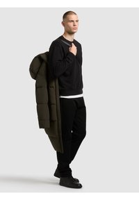 Czarna bluza, oliwkowa puchowa kurtka zarzucona na ramię, czarne spodnie i czarne sneakersy. Gładka faktura, casualowy design.
