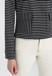 Czarna i biała bluzka w paski z długim rękawem, z dwoma przednimi kieszeniami i lekko dopasowanym fasonem. Noszona z jasnymi dżinsami.