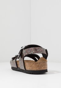 Błyszczące czarne sandały z regulowaną srebrną klamrą, korkową wkładką i teksturowaną podeszwą. Otwarta pięta i paski zapewniają przewiewną konstrukcję.