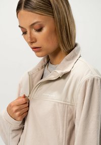 Tikkijakki vaaleassa beessissä, jossa on korkea kaulus ja puoleenväliin vetoketju. Ominaisuuksiin kuuluu teksturoitu kangas ja logo rinnassa. Sujuva vetoketjun nyöri.