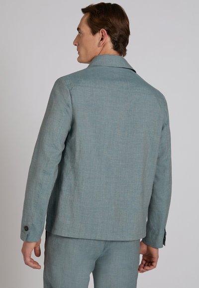 Mężczyzna o brązowych włosach ma na sobie jasnoniebieską marynarkę i dopasowane spodnie, pokazany od tyłu na jednolitym szarym tle.