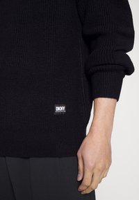 Pull en maille noire avec texture côtelée, arborant une étiquette logo DKNY. Les manches sont légèrement retroussées, mettant en valeur une coupe ajustée et une couleur unie.