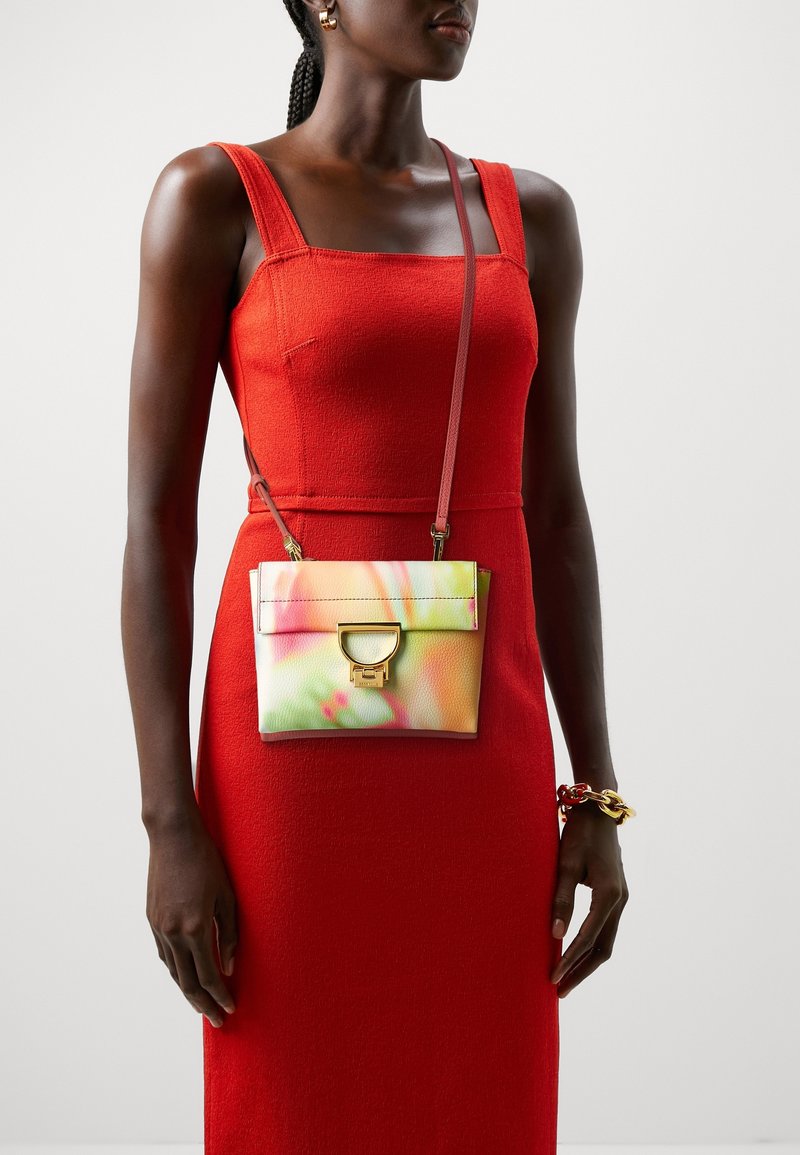 Femme portant une robe rouge sans manches, avec un petit sac bandoulière multicolore à boucle dorée et un bracelet épais à son poignet droit.