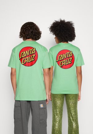 T-shirt in cotone verde con un grande logo circolare rosso "SANTA CRUZ" sulla schiena, indossato con pantaloni cargo grigi e pantaloni in paillettes verdi.