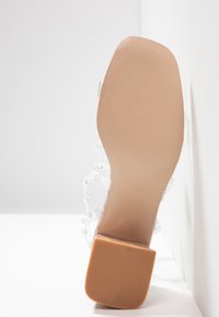 Sandale à talon avec une tige transparente et une semelle extérieure beige opaque. Dotée d'un talon carré et d'une semelle texturée pour une meilleure adhérence. Design minimaliste.