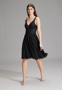 Robe noire avec un corsage à perles, décolleté en V et jupe en chiffon évasée. Modèle portant des sandales à talons argentées, se tenant contre un fond gris.