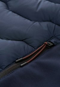 Tissu matelassé bleu marine avec un motif cousu ; présente une fermeture éclair noire et des cordons de traction rouges ; sections lisses et texturées.