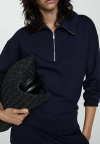 Sudadera azul marino con cuello de media cremallera, hecha de tela suave. Combinada con un bolso de mano negro de tejido y asas circulares.