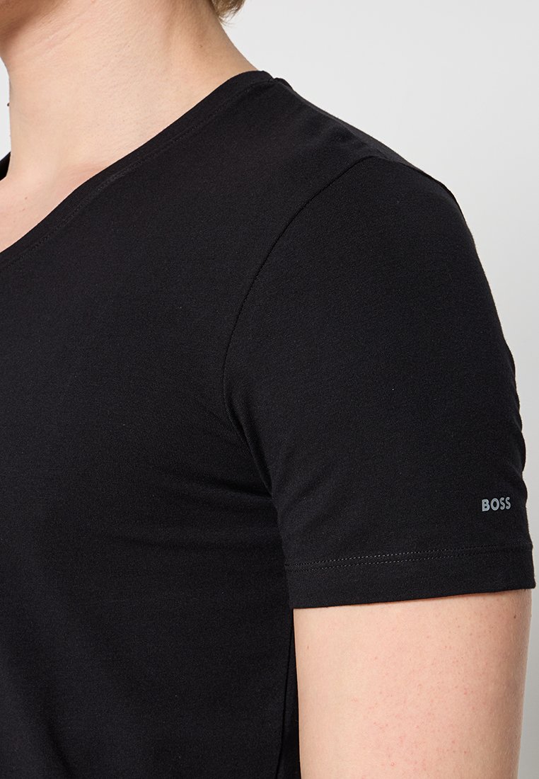 T-shirt noir à manches courtes en tissu doux, featuring un col en V et un petit logo sur la manche gauche. Texture lisse, ajusté.