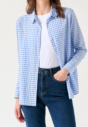 Femme portant une chemise à carreaux bleu clair et blanche boutonnée sur un top blanc, associée à un jean bleu foncé taille haute.