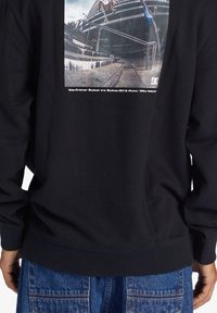 Felpa nera con collo tondo, caratterizzata da una stampa grafica rettangolare, polsini e orlo a costine. La grafica rappresenta una scena di skateboard.