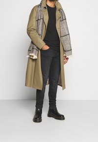 Trench de color verde oliva con una bufanda de cuadros grises, camiseta negra, jeans oscuros y botines Chelsea negros. Bufanda texturizada con flecos.