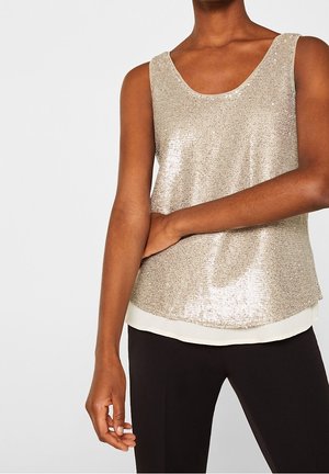 Femme portant un haut doré à sequins sans manches par-dessus une chemise blanche et un pantalon noir, avec un bras plié reposant sur sa taille.