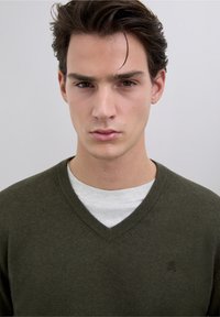 Suéter de cuello en v verde oscuro con puños y dobladillo acanalados, usado sobre una camiseta gris de cuello redondo. Textura suave con un pequeño logo en el pecho.