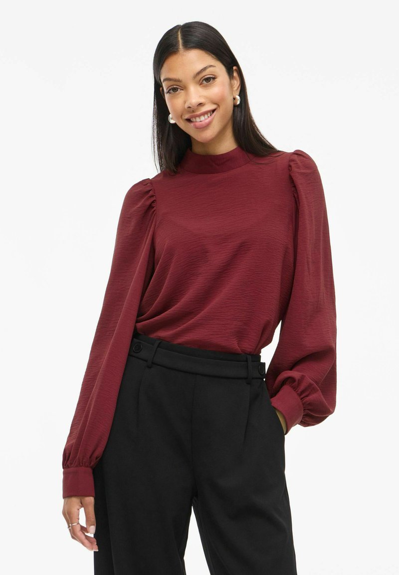 Blusa bordeaux con maniche lunghe, col alto e maniche a sbuffo, abbinata a pantaloni neri a vita alta con dettagli in bottoni.