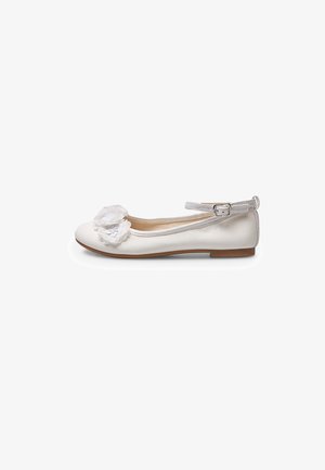 Zapato plano blanco con correa al tobillo y hebilla, decorado con dos flores de tela en capas en la parte delantera, suela marrón, vista lateral.