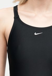 Κοντινό πλάνο ενός ατόμου που φοράει μαύρο αθλητικό τοπ Nike με λεπτές τιράντες και λευκό λογότυπο Nike στο στήθος.