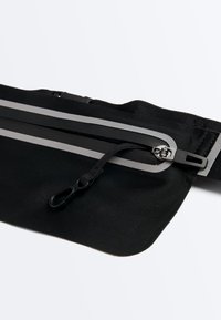Pochette de taille noire avec fermeture éclair réfléchissante et clip fixé sur une sangle noire, conçue pour un rangement portable sécurisé.