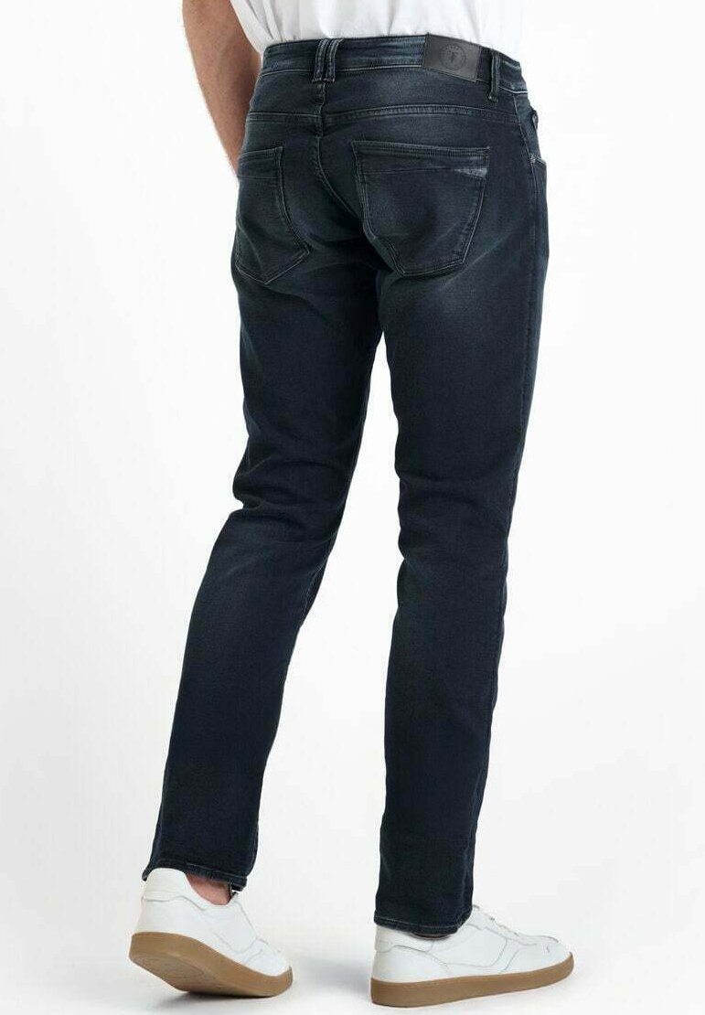 Personne portant un jean slim bleu foncé et des baskets blanches, vue de dos contre un fond blanc uni.
