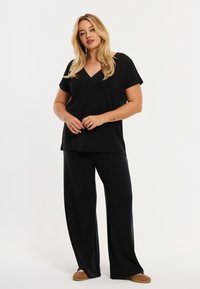 Sort V-hals top kombineret med løstsiddende sorte bukser. Stoffet virker blødt og let. Modellen står med hænderne foldet og viser et enkelt, afslappet design.