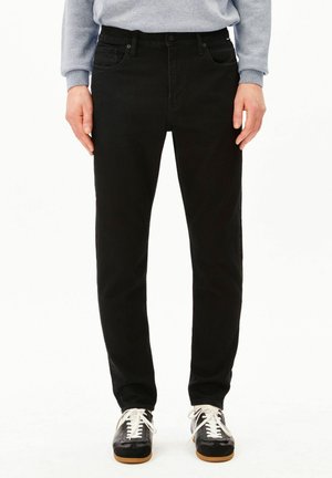 Personne portant un jean noir slim, un sweat-shirt gris clair et des baskets noires avec des lacets blancs, debout devant un fond blanc.