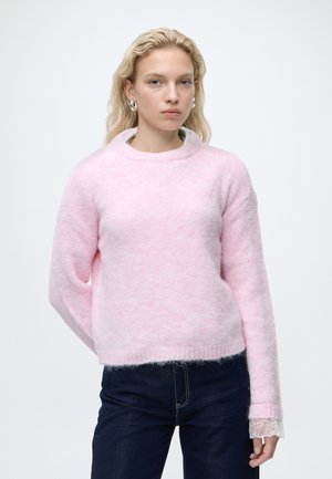 Femme blonde portant des boucles d'oreilles créoles en argent, un pull duveteux rose pâle, un jean foncé et un détail en dentelle visible au poignet sur fond blanc.
