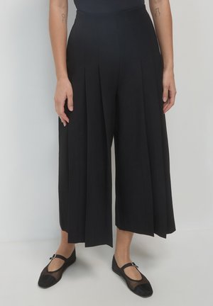 Schwarze weite Hosen mit Falten, hohem Bund und glatter Textur. Getragen mit schwarzen Ballerinas mit Mesh-Akzent und dekorativer Schnalle.