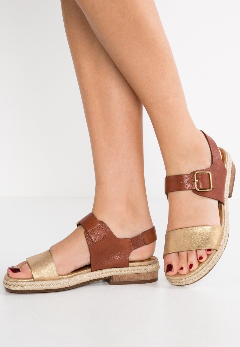 Sandales en cuir marron et doré avec une semelle de style espadrille, dotées d'une bride à boucle et d'un design à bout ouvert. Texture lisse avec un talon plat.