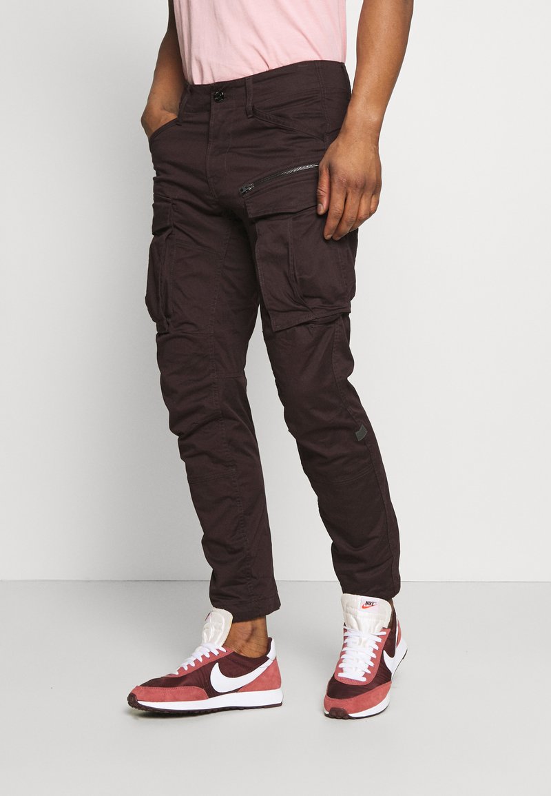 Pantaloni cargo de un maron închis, cu multiple buzunare, un buzunar lateral cu fermoar și o croială slim. Purtați cu adidași Nike roșii și albi.