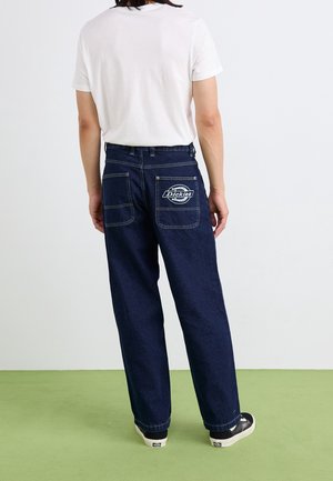 Personne portant un jean large bleu foncé avec le logo "Dickies" sur la poche arrière, un t-shirt blanc et des baskets noires, debout sur un sol vert.