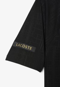 Svart kortärmad skjorta med ett strukturerat geometriskt mönster, som har en guld "LACOSTE"-logotyp på en svart tygetikett på ärmen.