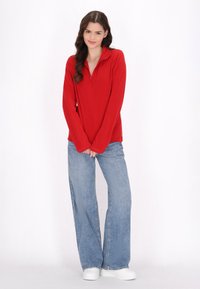 Rød pullover med lynlås krave, parret med lyseblå brede bukser. Stoffet ser blødt ud, og pasformen er afslappet. Hvide sneakers fuldender looket.