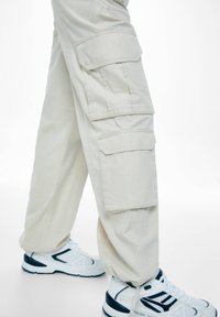 Pantalon cargo beige avec plusieurs poches latérales, légère bande élastique aux chevilles, associé à des chaussures de sport blanches avec des accents bleus.