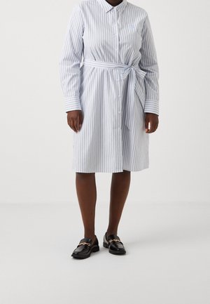 Personne portant une robe blanche jusqu'aux genoux avec de fines rayures verticales et une ceinture nouée, associée à des mocassins noirs avec des détails en chaîne dorée.