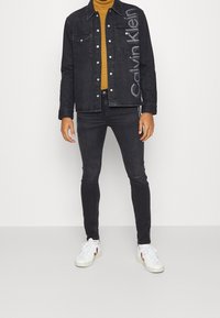 Chaqueta de mezclilla negra con cierre de botones y dos bolsillos frontales. Lleva una camiseta de cuello alto mostaza y se combina con jeans negros ajustados y zapatillas blancas.