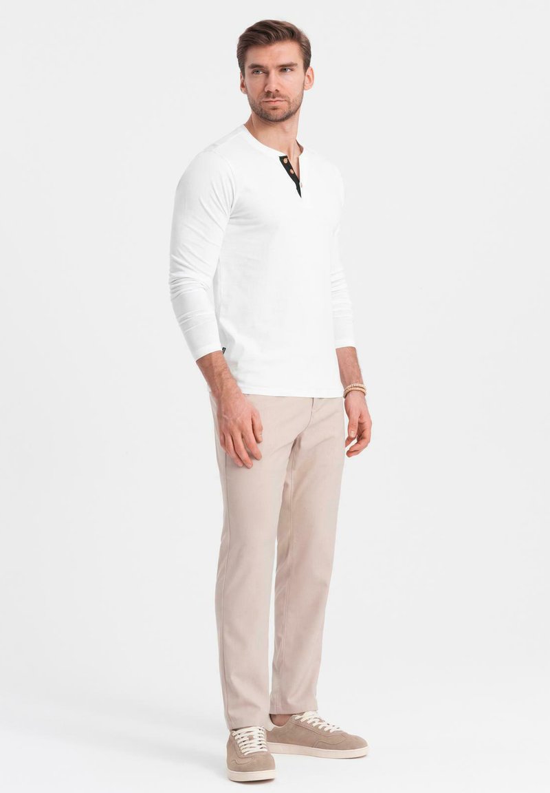 Camicetta bianca a maniche lunghe modello henley con bottoni neri, abbinata a pantaloni chiari beige e sneakers color nocciola, su uno sfondo neutro.