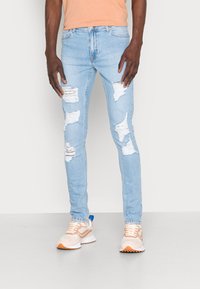 Light blue ripped jeans with distressed patches and a slim fit. Paired with beige sneakers featuring a blue accent and a white sole.