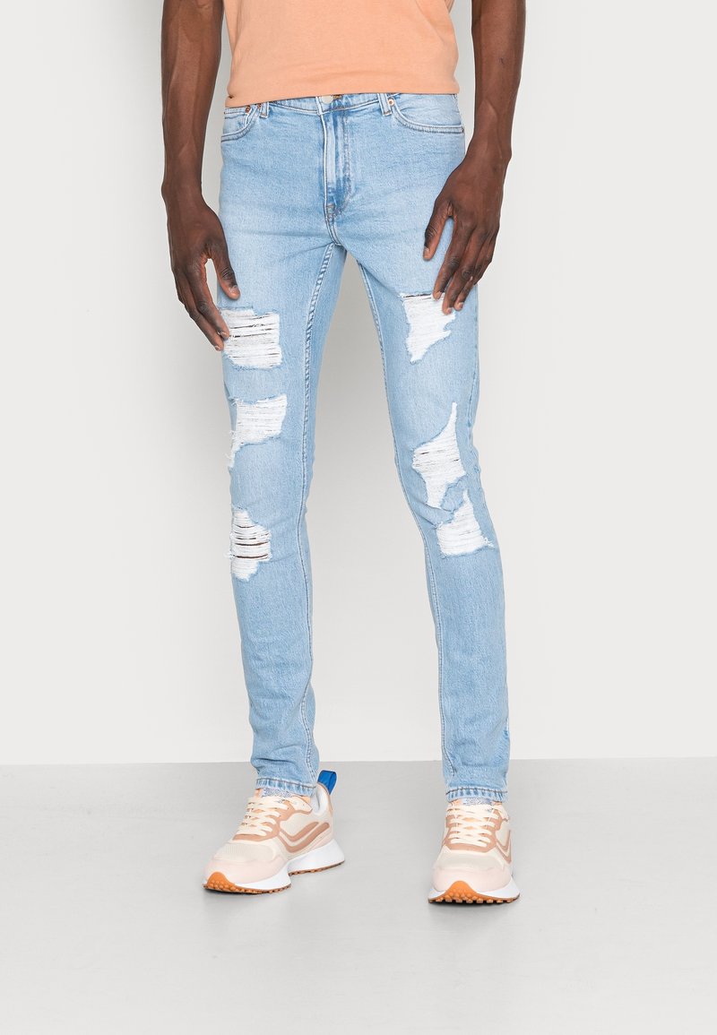Light blue ripped jeans with distressed patches and a slim fit. Paired with beige sneakers featuring a blue accent and a white sole.