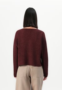 Femme avec des cheveux noirs raides à longueur d'épaule portant un pull long à manches longues rouge foncé et un pantalon beige, se tenant face à un mur blanc.