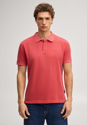 Junger Mann mit lockigem Haar, der ein korallenfarbenes Polohemd und blaue Jeans trägt, steht vor einem schlichten hellen Hintergrund.