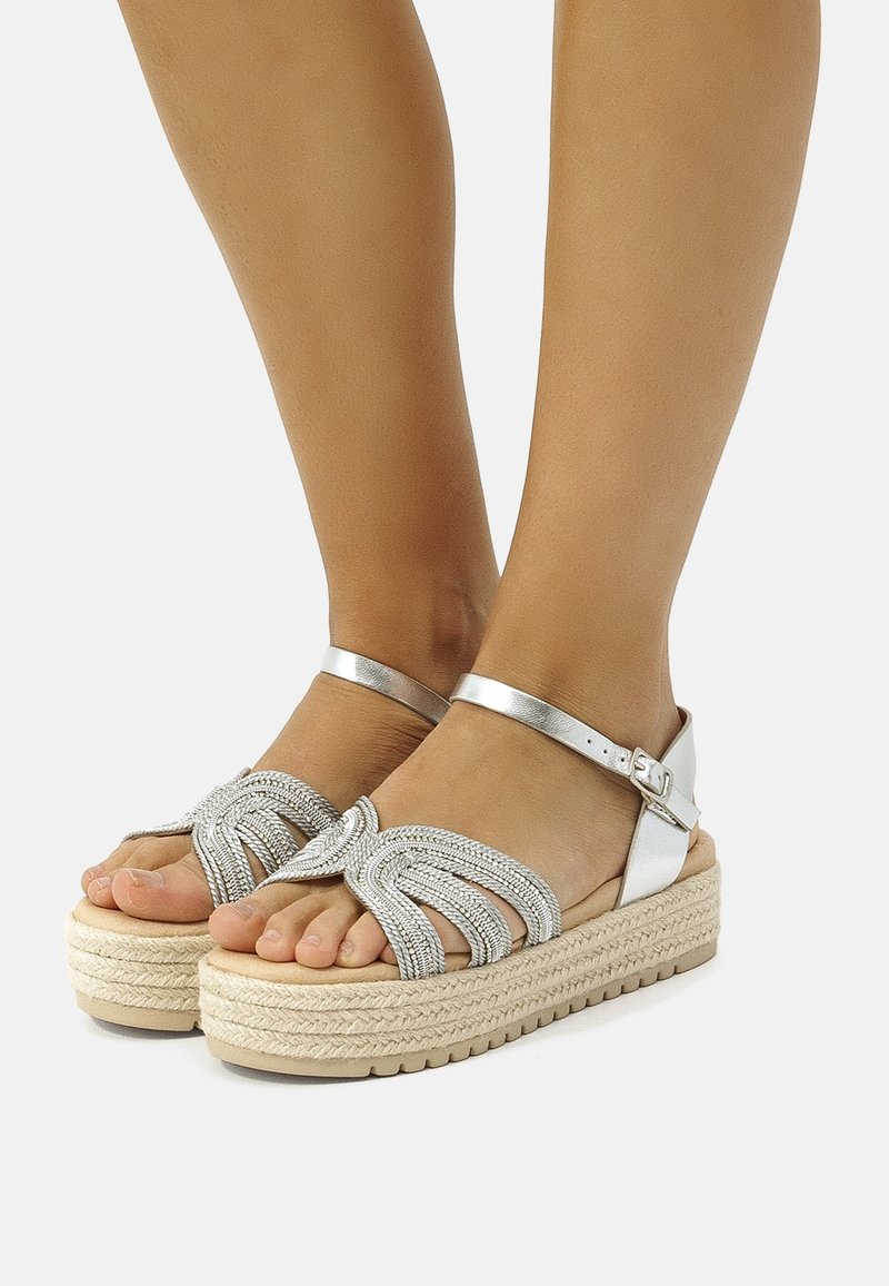 Sandales plateformes argentées avec un design supérieur tressé, une bride de cheville ajustable et une semelle en jute tissée. Matériau lisse semblable au cuir et texture légère.
