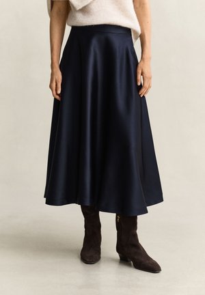 Femme portant une jupe midi en satin bleu marine, un pull beige et des bottines en daim marron foncé, debout sur un sol clair avec un fond neutre.