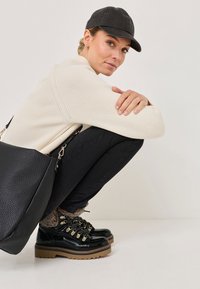 Fekete lakkbőr csizmák arany részletekkel, bézs kötött pulóver és fekete sapka. Egy texturált fekete táska fekszik a guggoló alak mellett.