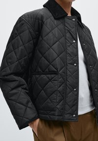 Zwarte gewatteerde jas met een ribfluwelen kraag, drukknoopsluitingen en een zijzak. De textuur is glad met een gewatteerd ontwerp voor extra warmte.