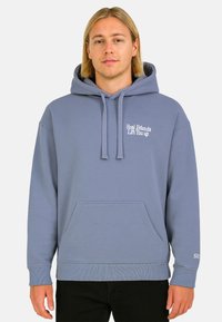 Young man with shoulder-length blonde hair wearing a light blue hoodie with the text "Real Friends Lift You up" on the chest.