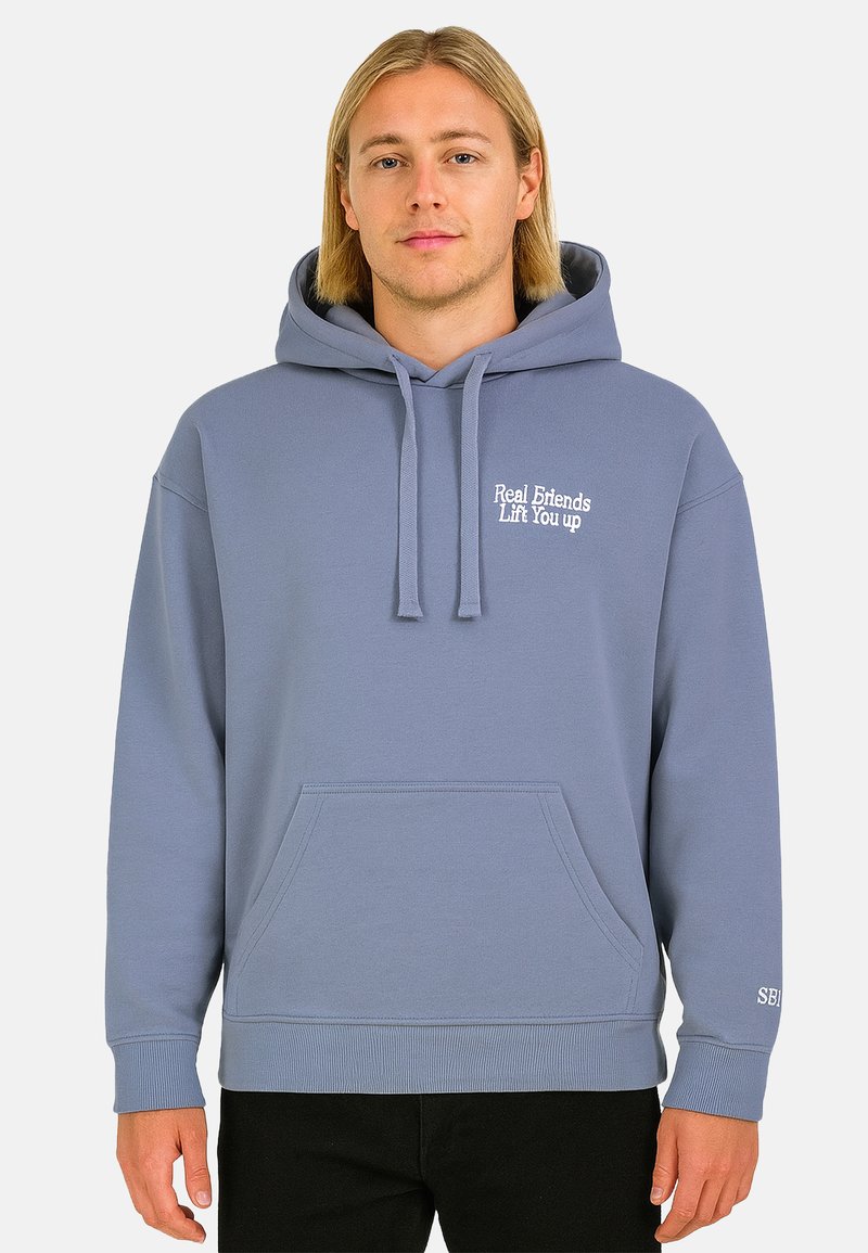 Young man with shoulder-length blonde hair wearing a light blue hoodie with the text "Real Friends Lift You up" on the chest.